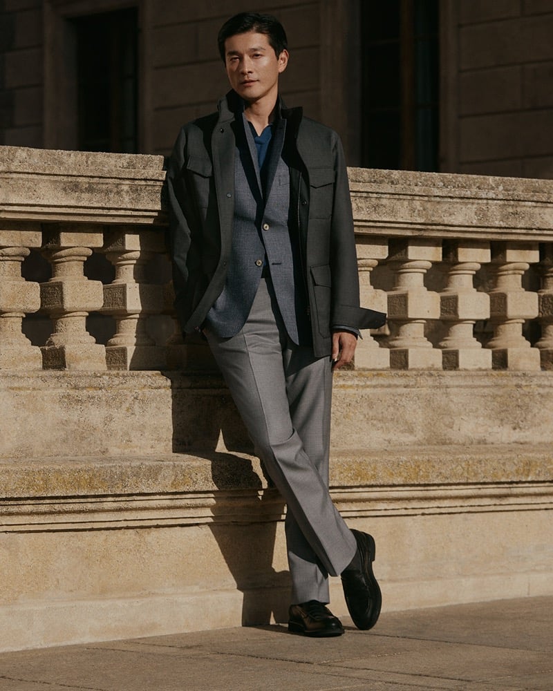 Daisuke leans against a carved stone balustrade in a field jacket layered over a blue blazer with gray trousers and black loafers, for Canali's spring-summer 2026 campaign
