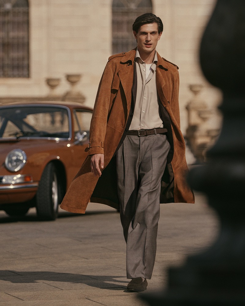 Edoardo walks down a European city street past a vintage orange sports car in a long brown suede trench, cream shirt, and gray pleated trousers for Canali's spring-summer 2026 campaign