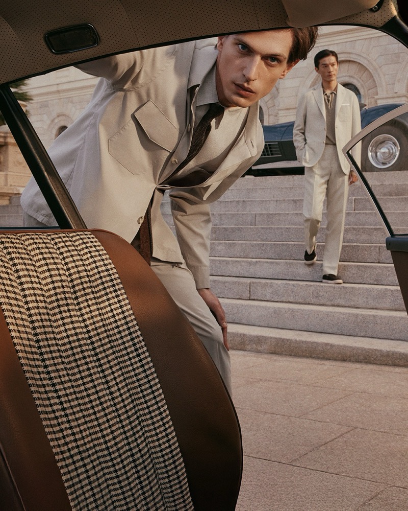Edoardo leans into the open door of a vintage car in a beige safari-style suit and brown knit tie while Daisuke stands on stone steps beyond in a cream linen suit and dark tie for Canali's spring-summer 2026 campaign