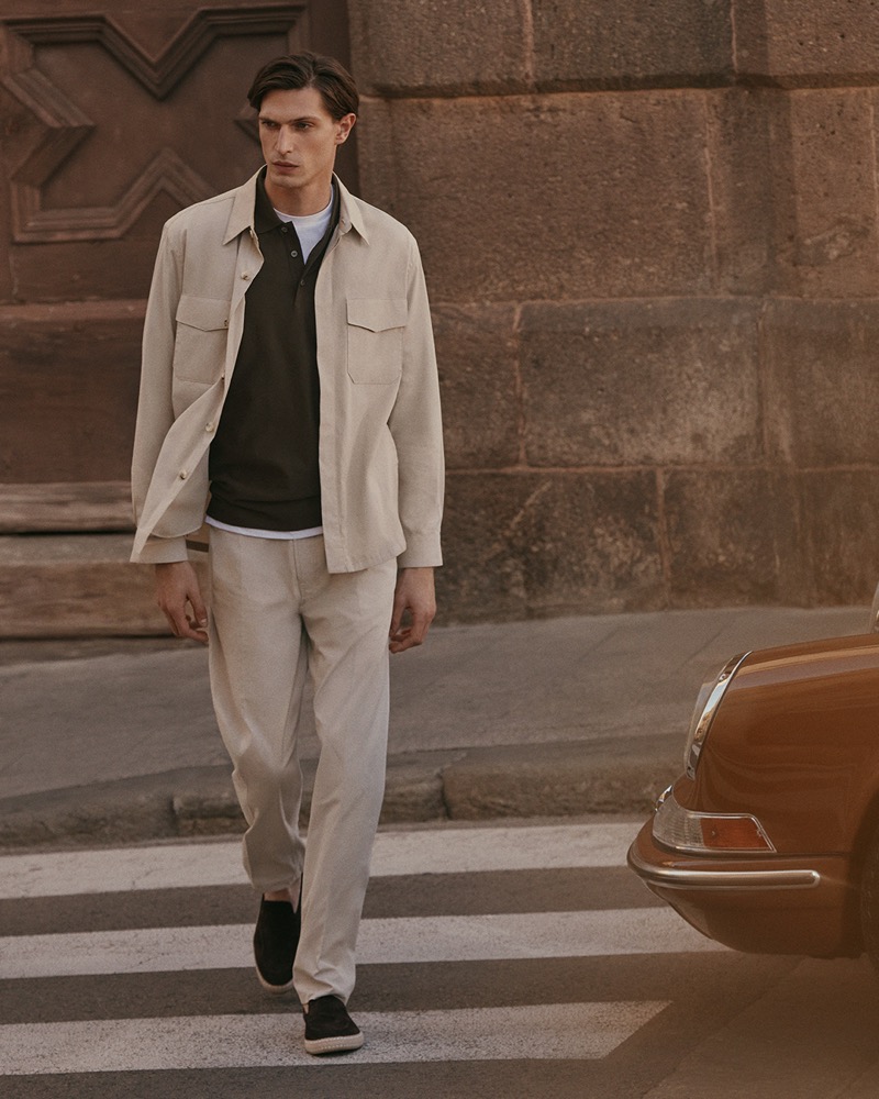 Edoardo crosses a street on a crosswalk past an orange vintage sports car in a linen overshirt over a dark brown knit polo, cream trousers, and dark espadrilles for Canali's spring-summer 2026 campaign