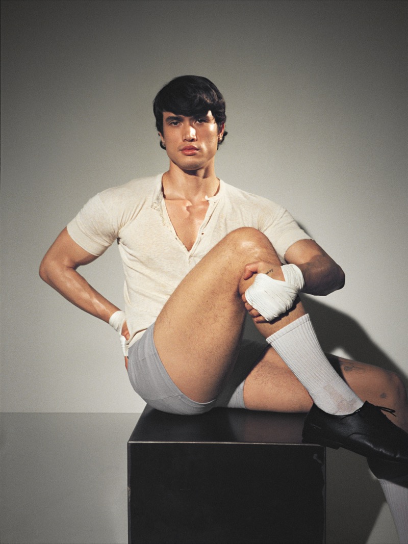 Melton sits on a dark cube in a cream henley, gray shorts, white tube socks, and black leather shoes with wrapped hands