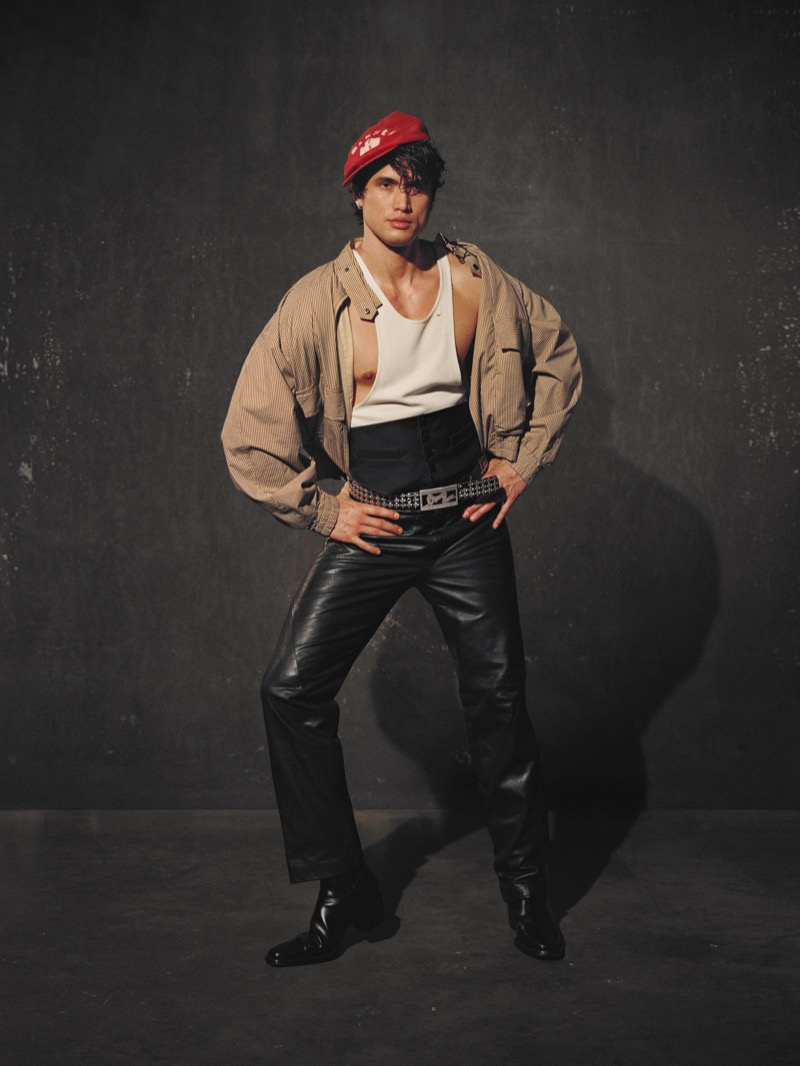 Melton stands in a tan striped jacket over a white tank top with a black cummerbund, leather pants, studded belt, and red beret