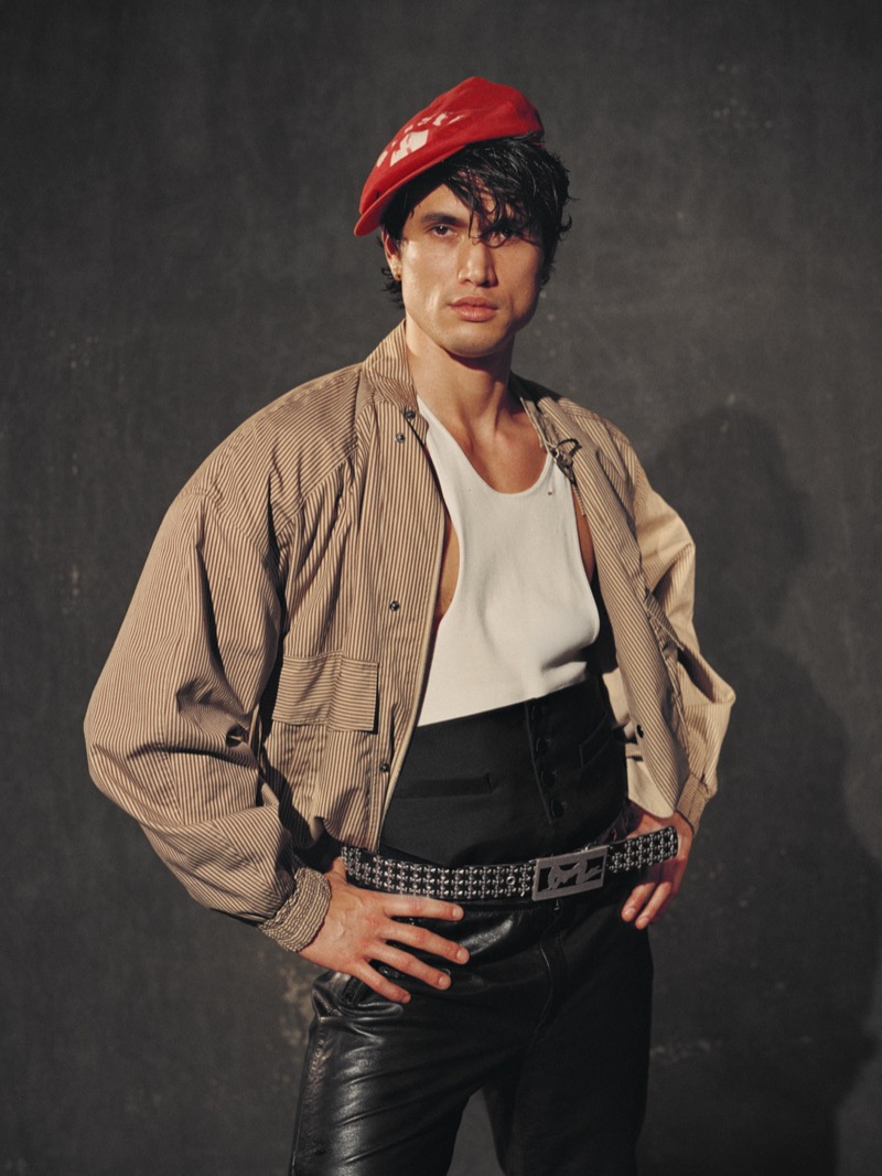 Melton in a tan striped jacket over a white tank top with leather pants and a red beret in a half-body shot