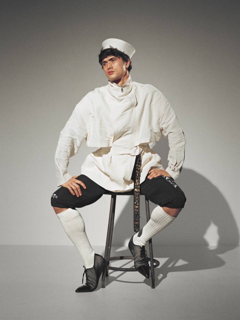 Melton sits on a stool in a white belted jacket, black shorts, knee socks, and a white pillbox hat