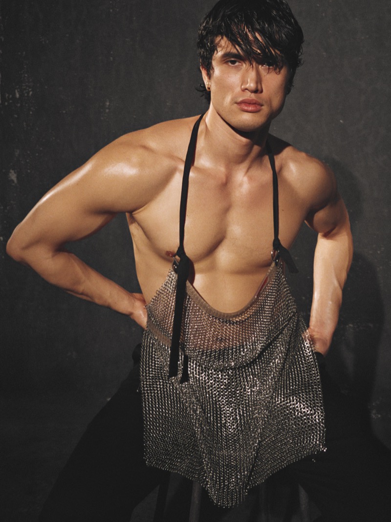 Charles Melton stands shirtless wearing a silver chainmail halter top with oiled skin