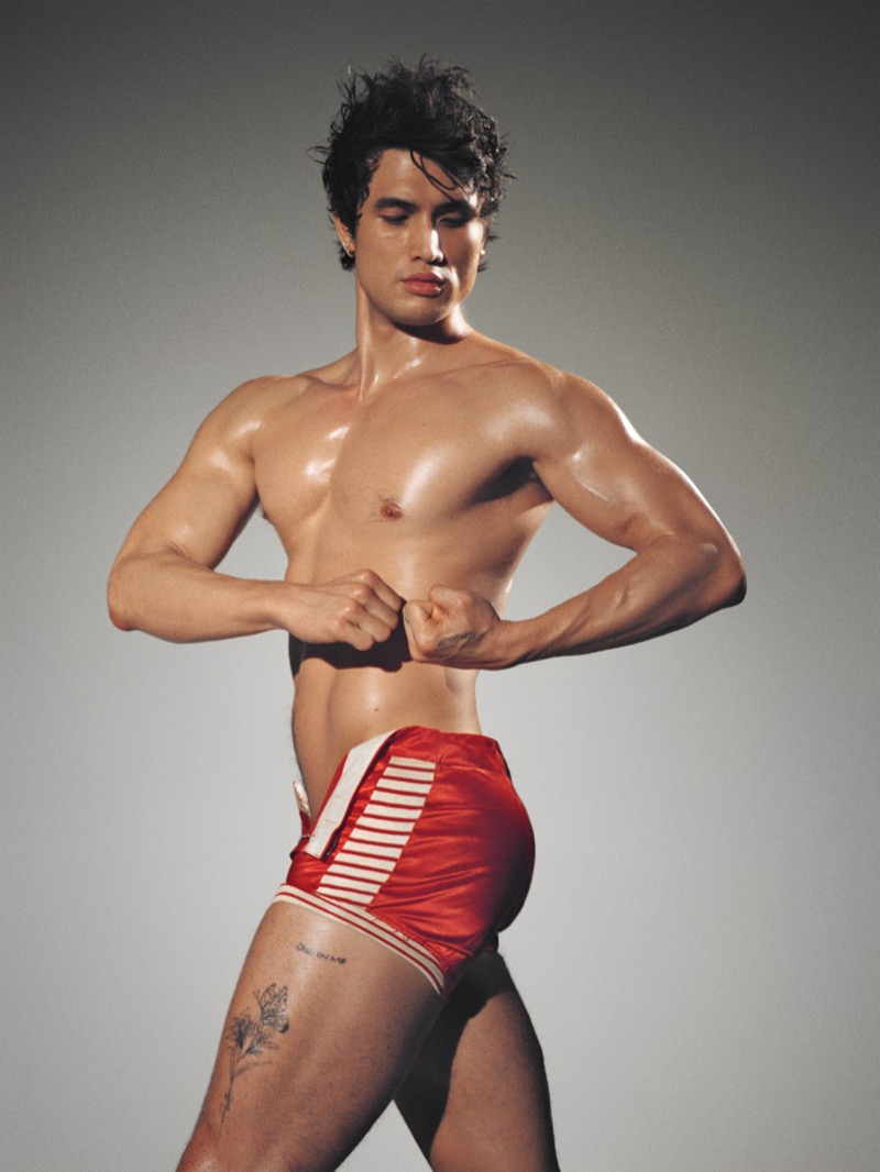 Charles Melton strikes a bodybuilder pose in red athletic shorts with white striped trim and oiled skin