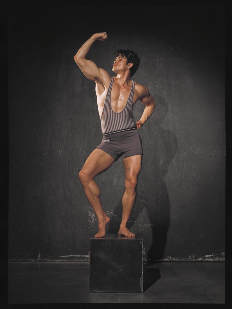 Charles Melton flexes in a striped halter bodysuit and gray shorts with one foot on a dark cube