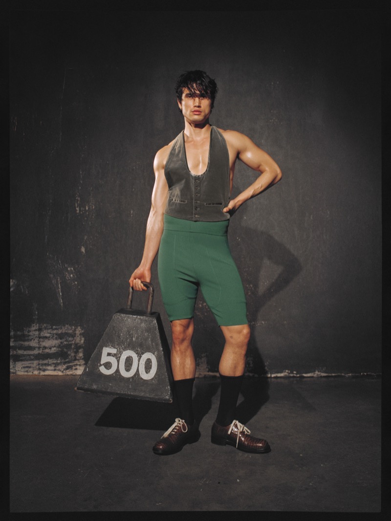 Charles Melton holds a 500-pound weight prop in a gray vest and green cycling shorts with black socks