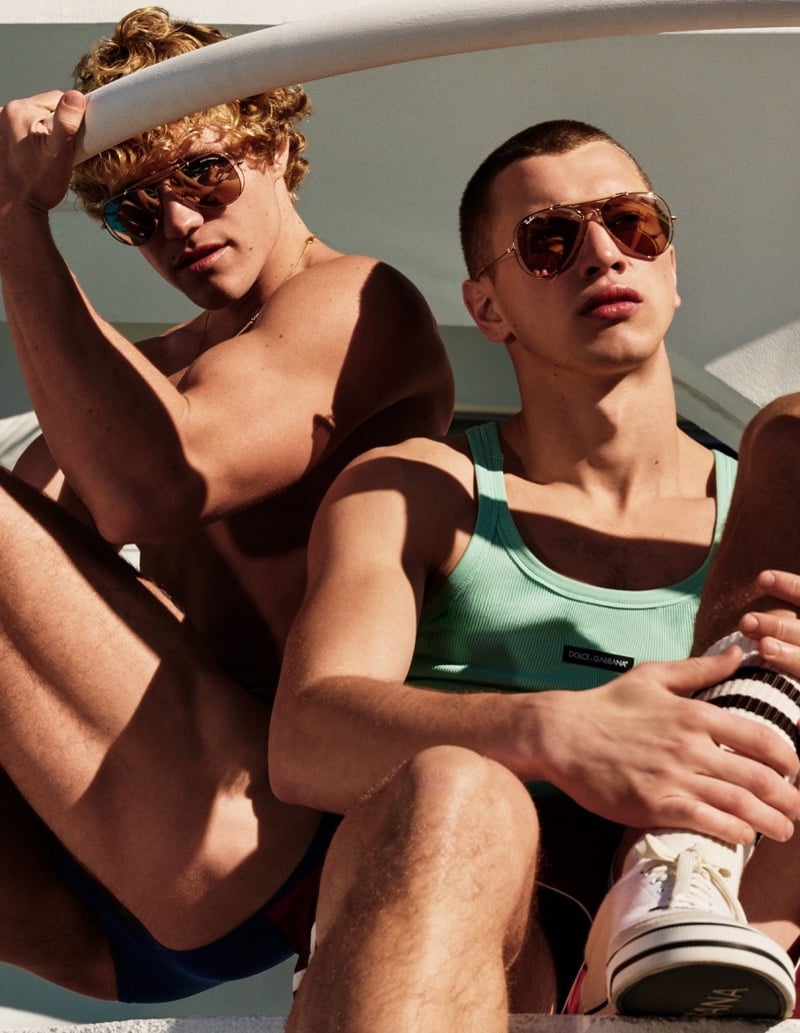 Two models poolside in Dolce & Gabbana x Ray-Ban Outdoorsman II aviators, one in blue swim shorts and one in a mint Dolce & Gabbana tank top