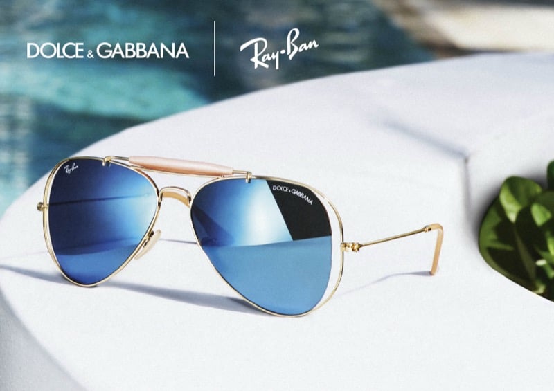 Gold Dolce & Gabbana x Ray-Ban Outdoorsman II aviators with mirrored blue lenses and a mother-of-pearl brow bar resting on a white surface beside a pool
