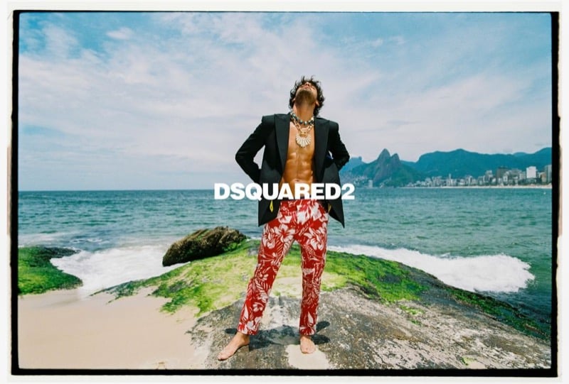 Marlon stands barefoot on a rock at Ipanema Beach in a black blazer over a bare chest, red and white hibiscus print trousers, and layered jewelry, with the Two Brothers mountains behind him, for Dsquared2's spring-summer 2026 campaign