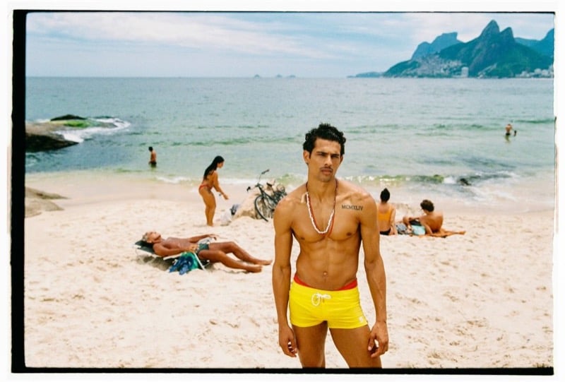 Jonas Barros stands shirtless on the sand at Ipanema Beach in yellow swim boxers and a beaded necklace, with the Two Brothers mountains in the distance, for Dsquared2's spring-summer 2026 campaign