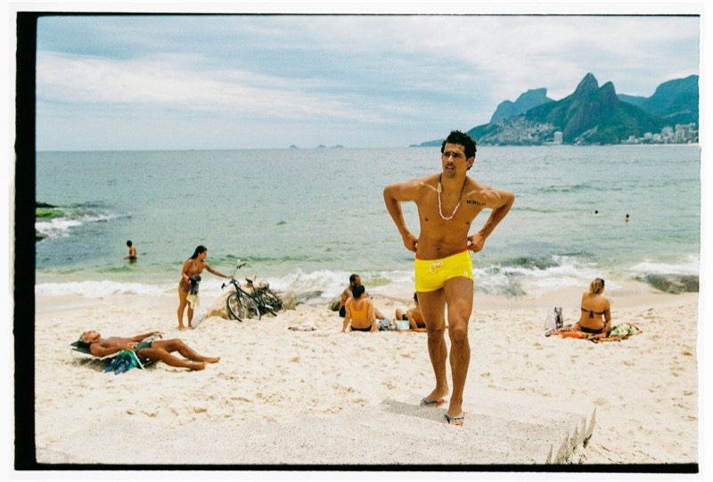 Jonas Barros stands shirtless on Ipanema Beach with hands on hips in yellow swim trunks, with sunbathers and the Two Brothers mountains behind him, for Dsquared2's spring-summer 2026 campaign