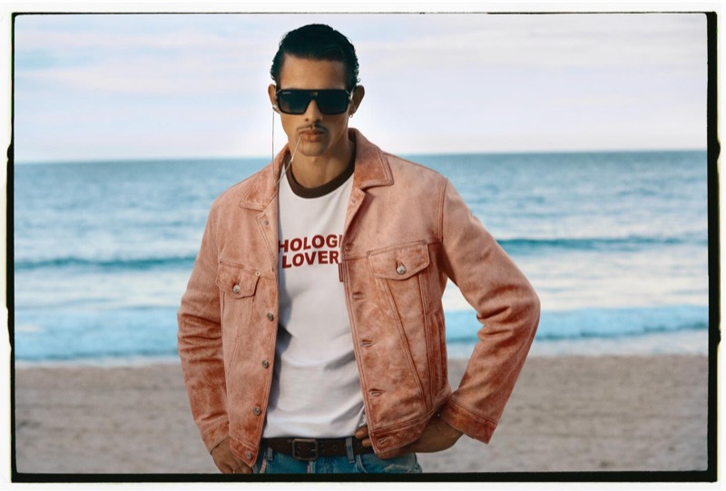 Jonas stands on the beach in a red cracked leather jacket over a white "Pathological Lover" graphic tee and dark sunglasses for Dsquared2's spring-summer 2026 campaign