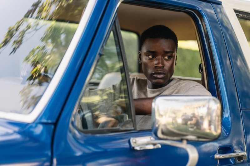 Babacar sits behind the wheel of a blue pickup truck in a heather grey sweatshirt
