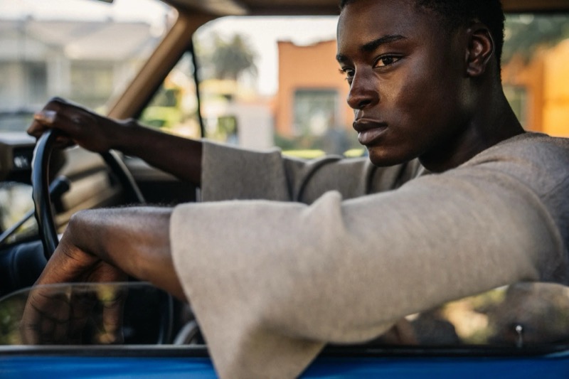 Babacar rests his arms on the steering wheel of a blue truck in a heather grey long-sleeve top