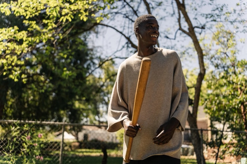 Babacar stands in a backyard holding a bat in a sweatshirt and dark trousers