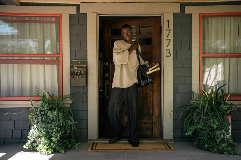 Babacar stands in a doorway with a bag and bat in an long-sleeve top and dark trousers