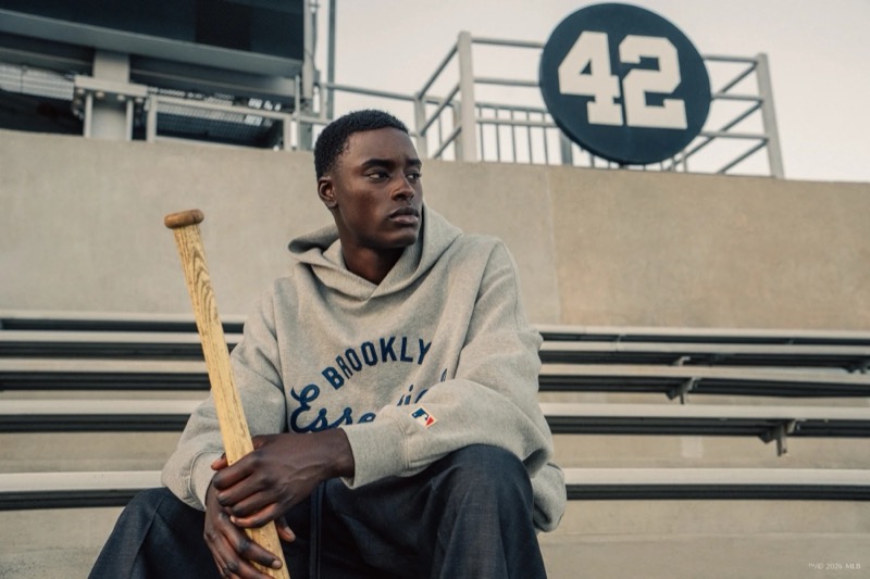Babacar sits on bleachers holding a bat in a Brooklyn Essentials hoodie with the number 42 behind him