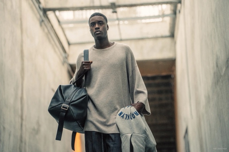 Babacar walks through a concrete corridor carrying a leather bag and a Brooklyn hoodie in a heather grey sweatshirt