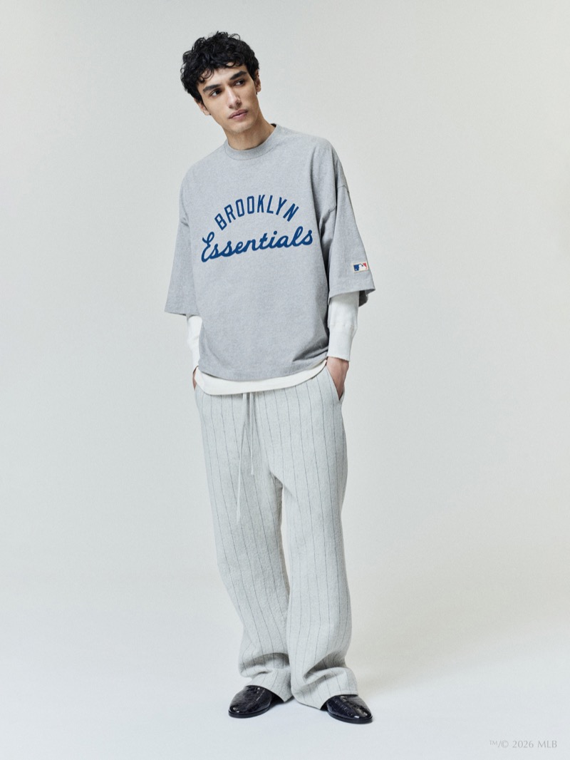 A model wears the Fear of God Essentials Jackie Robinson Day merch tee with Brooklyn lettering over pinstripe trousers