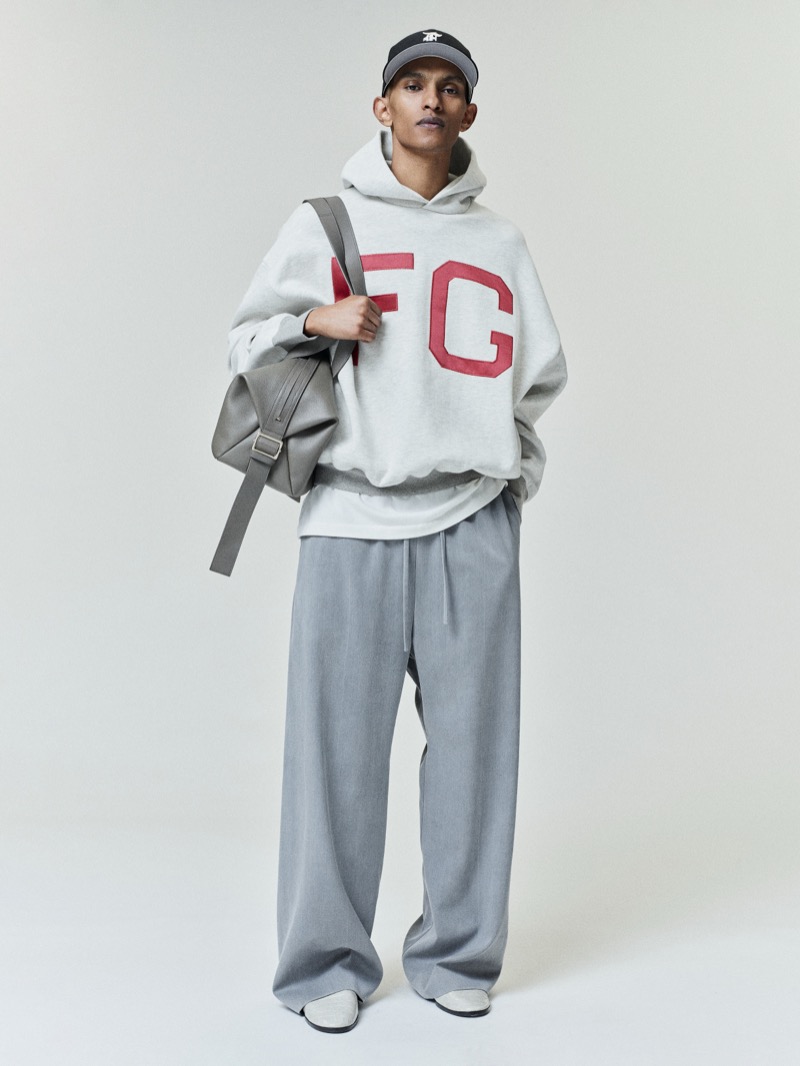 A model wears the Fear of God Essentials NLBM sport hoodie with red FG lettering, grey drawstring trousers, a baseball cap, and a leather bag