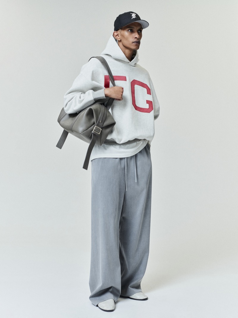 A model turns to the side in the Fear of God Essentials NLBM sport hoodie with red FG lettering, a baseball cap, grey trousers, and a leather bag