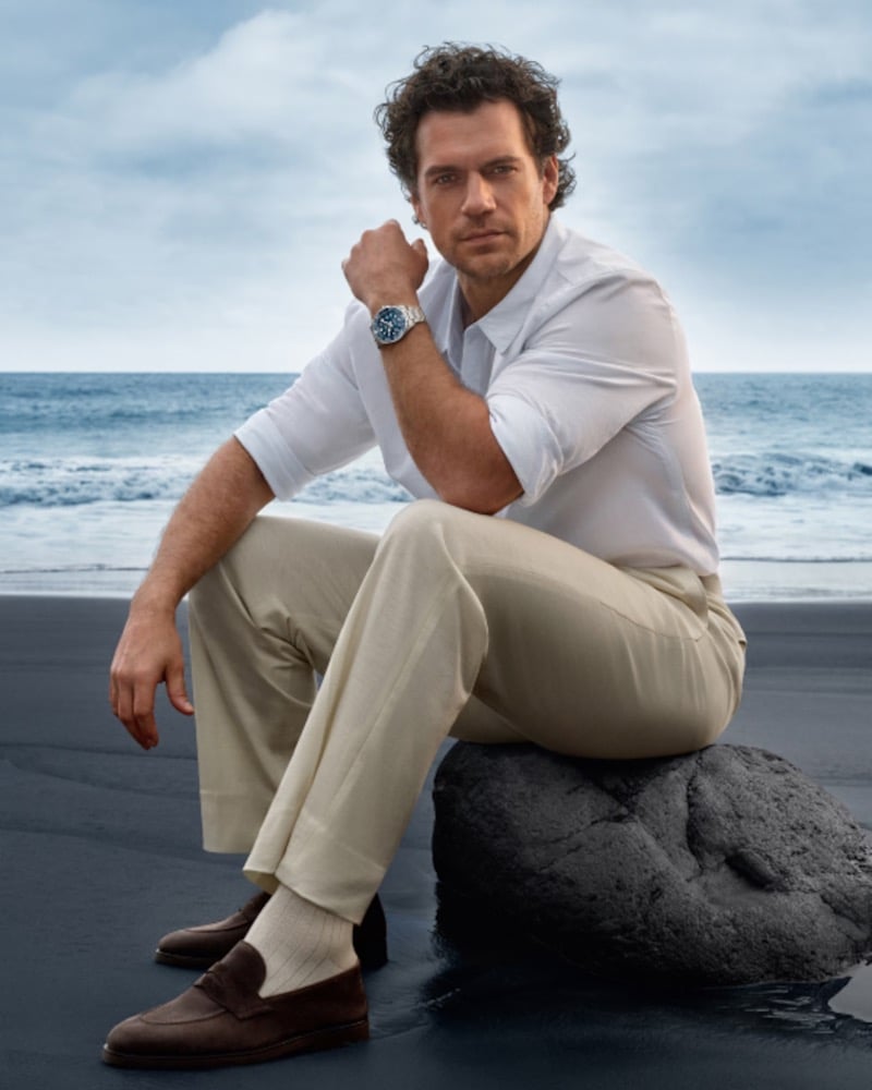 Cavill sits on a rock at the shore wearing a white shirt and the Longines HydroConquest
