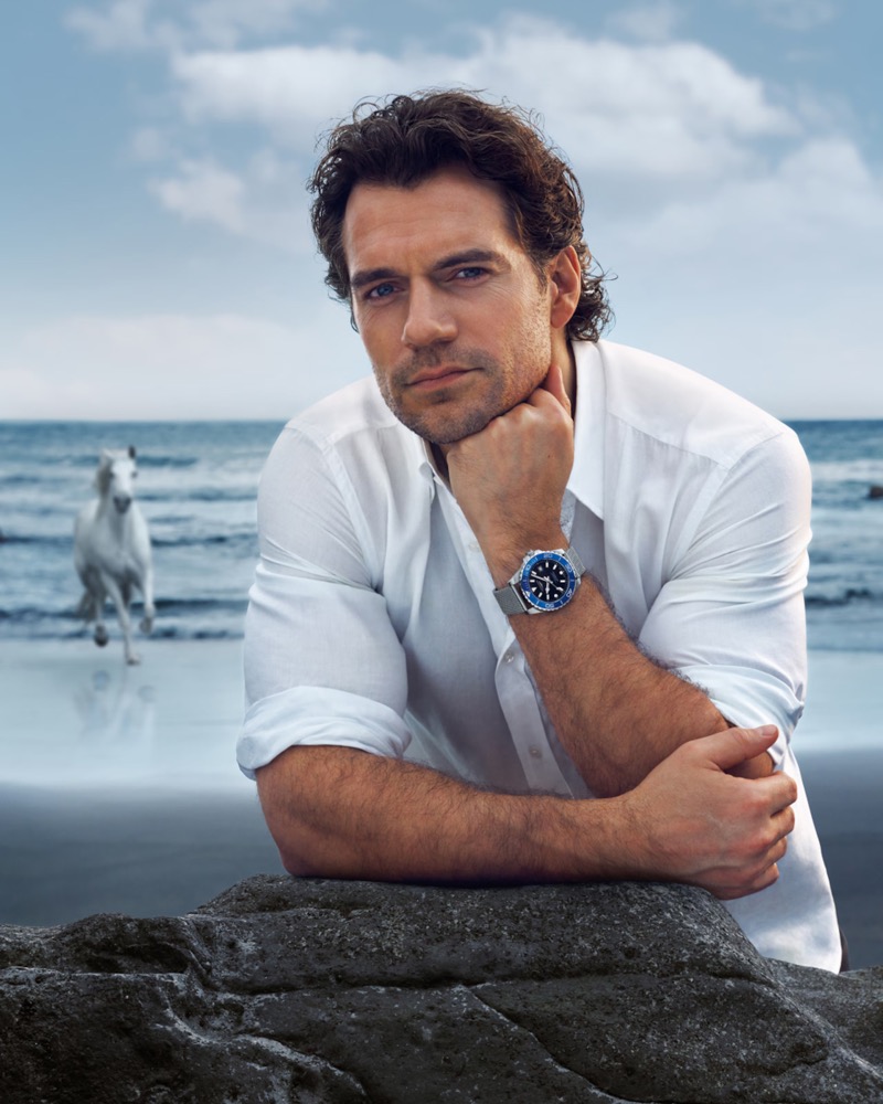 Cavill leans on a rock in a white shirt wearing the Longines HydroConquest with a blue ceramic bezel