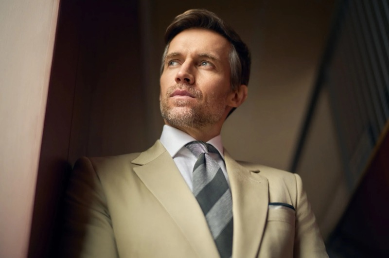 Close-up of Andrew in a tan one-button suit with a pale striped shirt, a gray striped tie, and a pocket square, standing in warm interior light for Huntsman's spring-summer 2026 collection