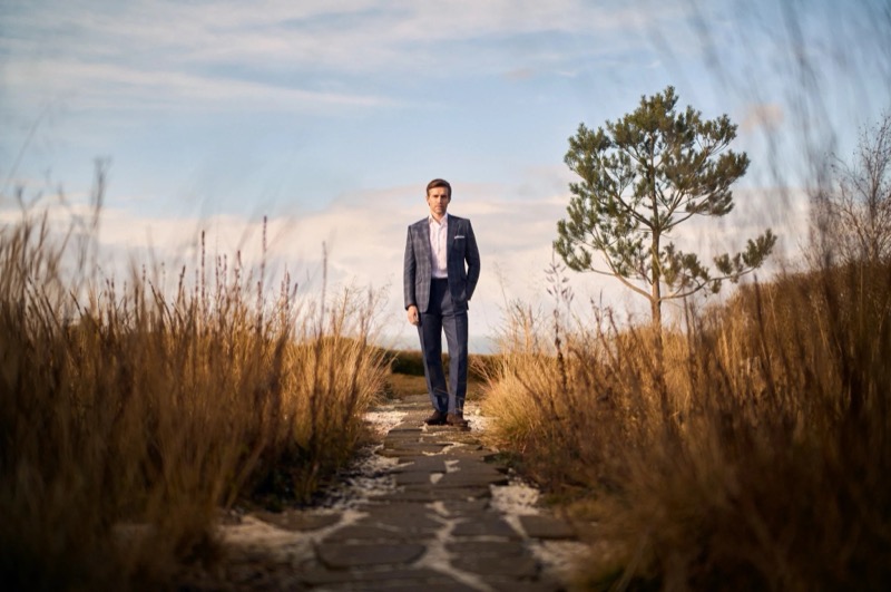 Andrew stands on a stone path through tall coastal grass in a navy check one-button suit, pale pink shirt, and brown shoes under an open sky for Huntsman's spring-summer 2026 collection
