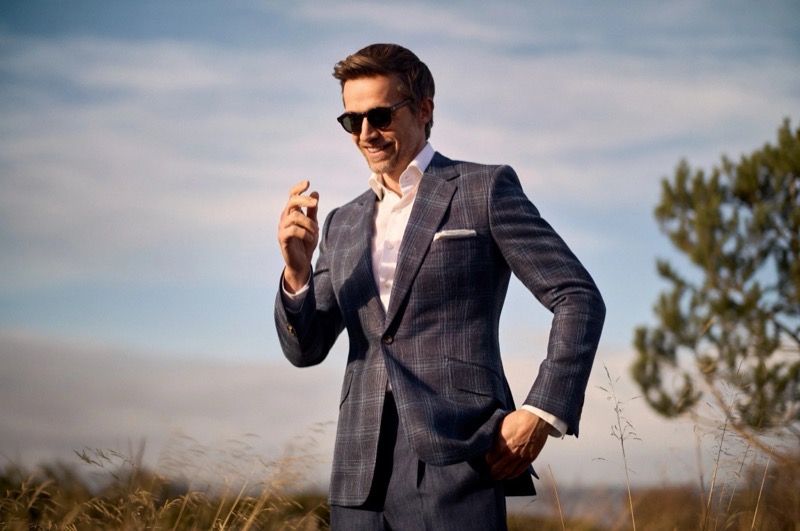 Andrew laughs in a blue windowpane plaid one-button suit worn with an open white shirt and dark sunglasses, standing in tall grass under a clear sky for Huntsman's spring-summer 2026 collection