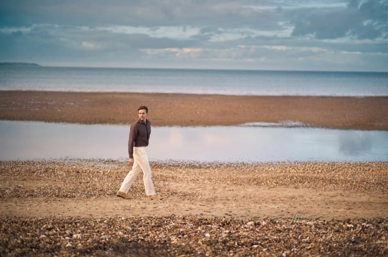 Andrew walks the shingle at low tide in a brown long-sleeve shirt and cream wide-leg trousers with the sea and cloud behind him for Huntsman's spring-summer 2026 collection