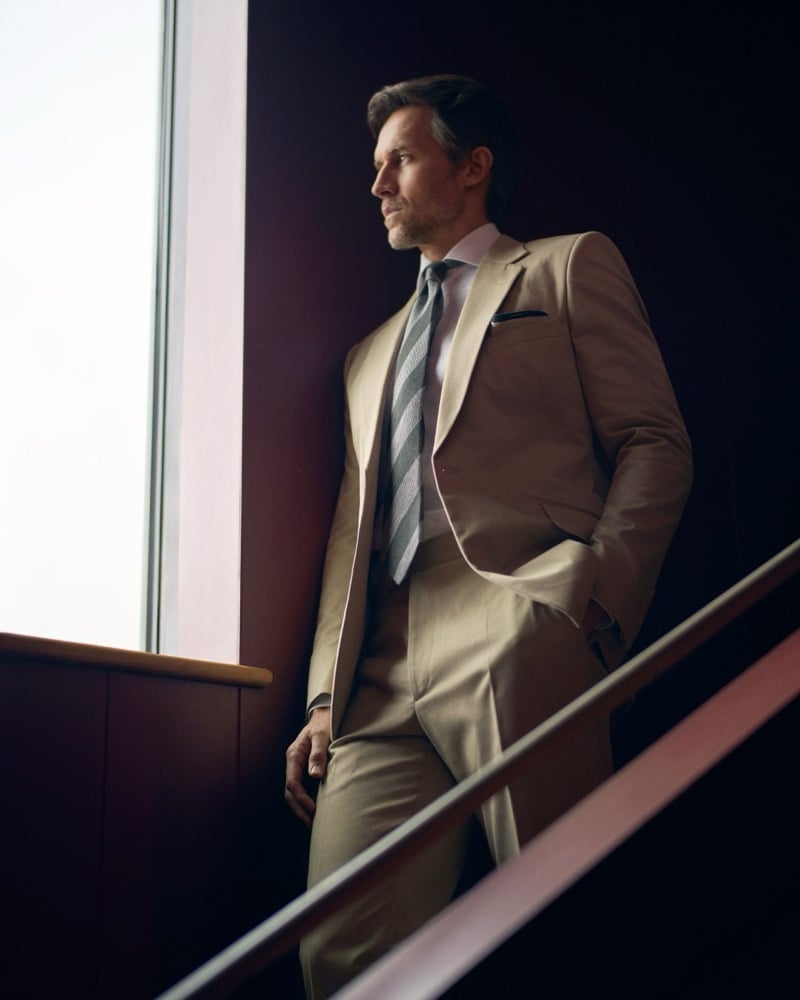 Andrew stands on a staircase beside a maroon wall in a tan one-button suit, pink shirt, gray striped tie, and pocket square for Huntsman's spring-summer 2026 collection