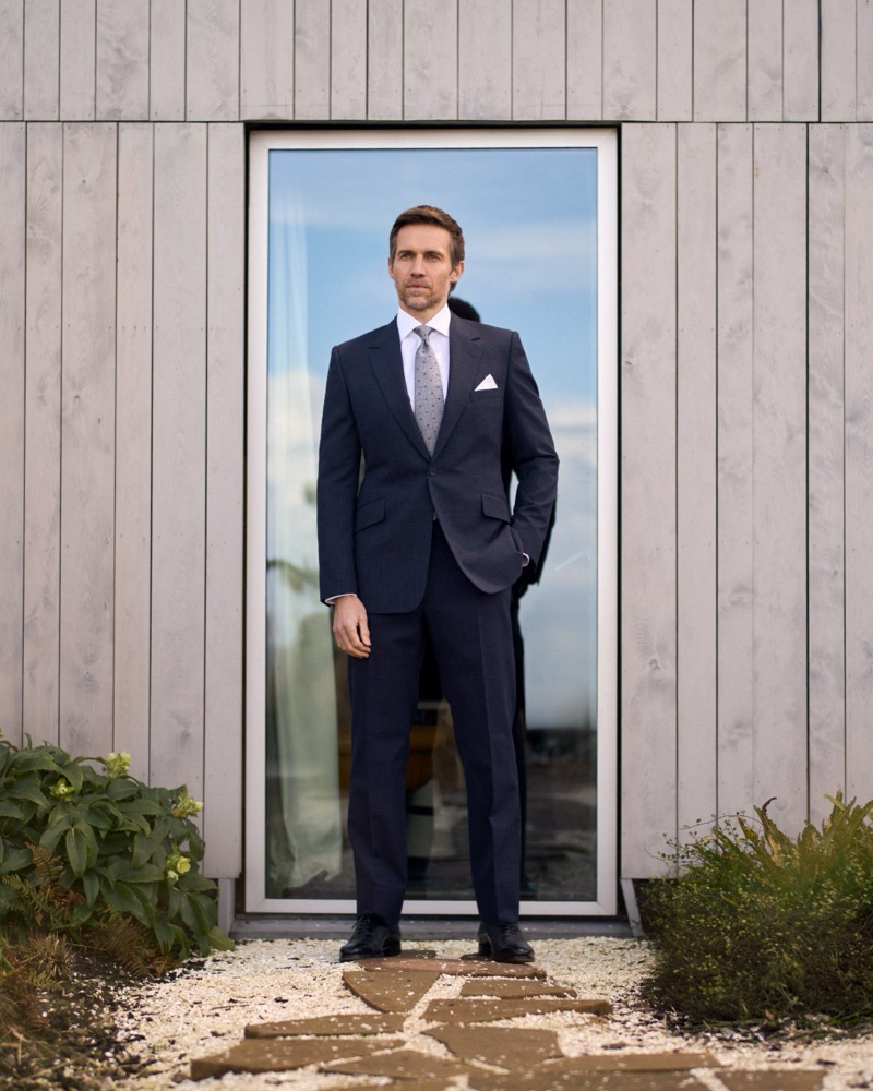 Andrew stands in the doorway of a modern wood-clad coastal house in a navy one-button suit, white shirt, dotted tie, and pocket square with stone paving underfoot for Huntsman's spring-summer 2026 collection