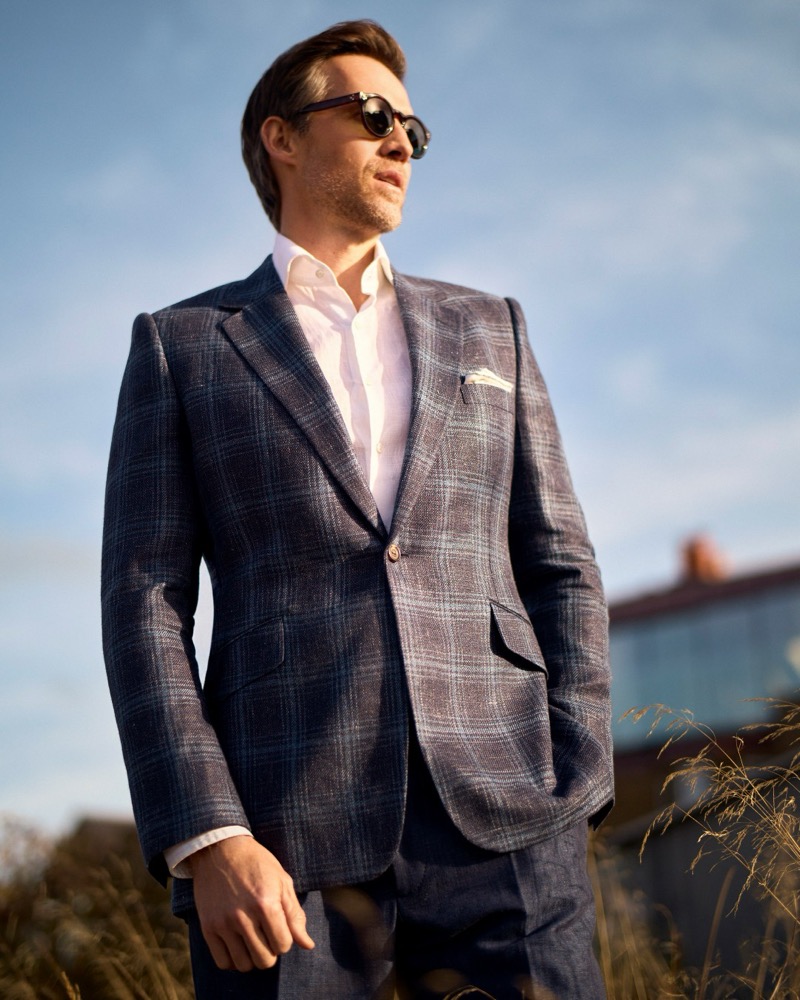 Andrew wears a navy and brown plaid one-button suit, a white shirt, and dark round sunglasses with a pocket square, standing in golden-hour coastal grass for Huntsman's spring-summer 2026 collection