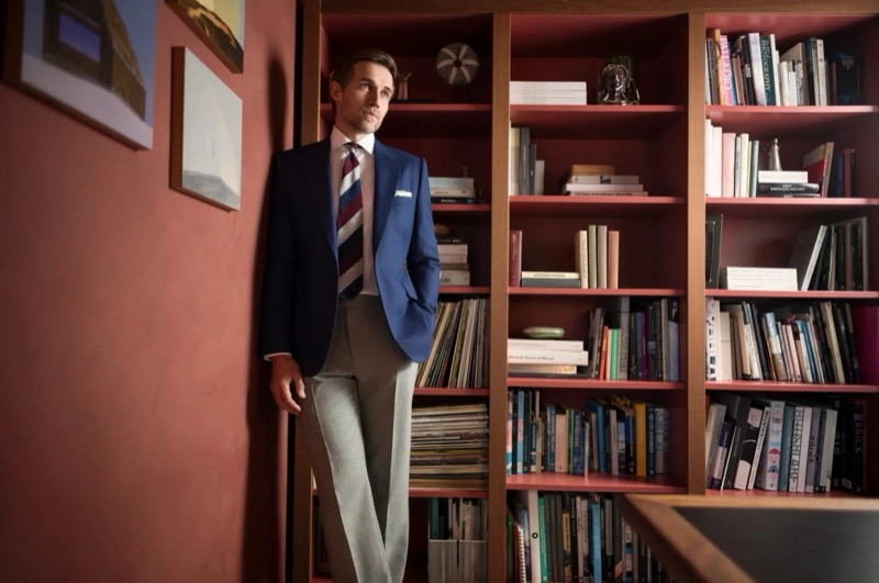 Andrew leans against a red-walled library shelf in a blue one-button blazer, gray trousers, white shirt, and a striped navy and burgundy tie for Huntsman's spring-summer 2026 collection