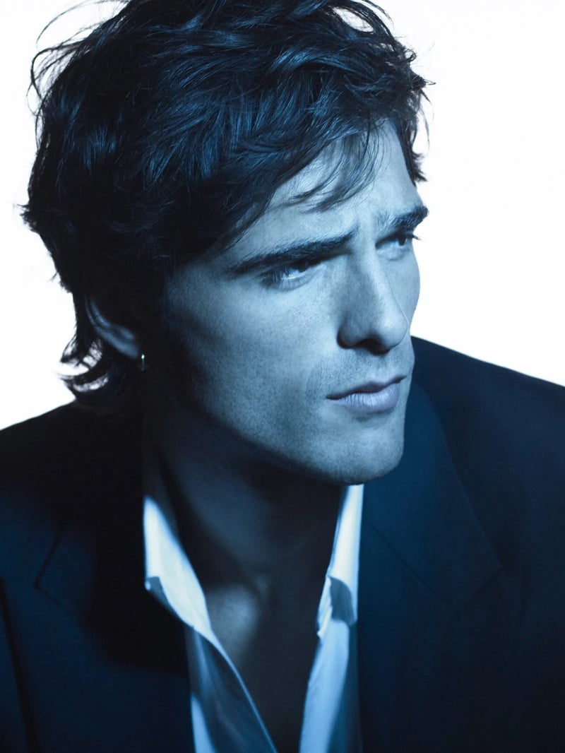 Blue-toned three-quarter portrait of Elordi in a dark suit jacket and open white shirt with a small hoop earring for Bleu de Chanel