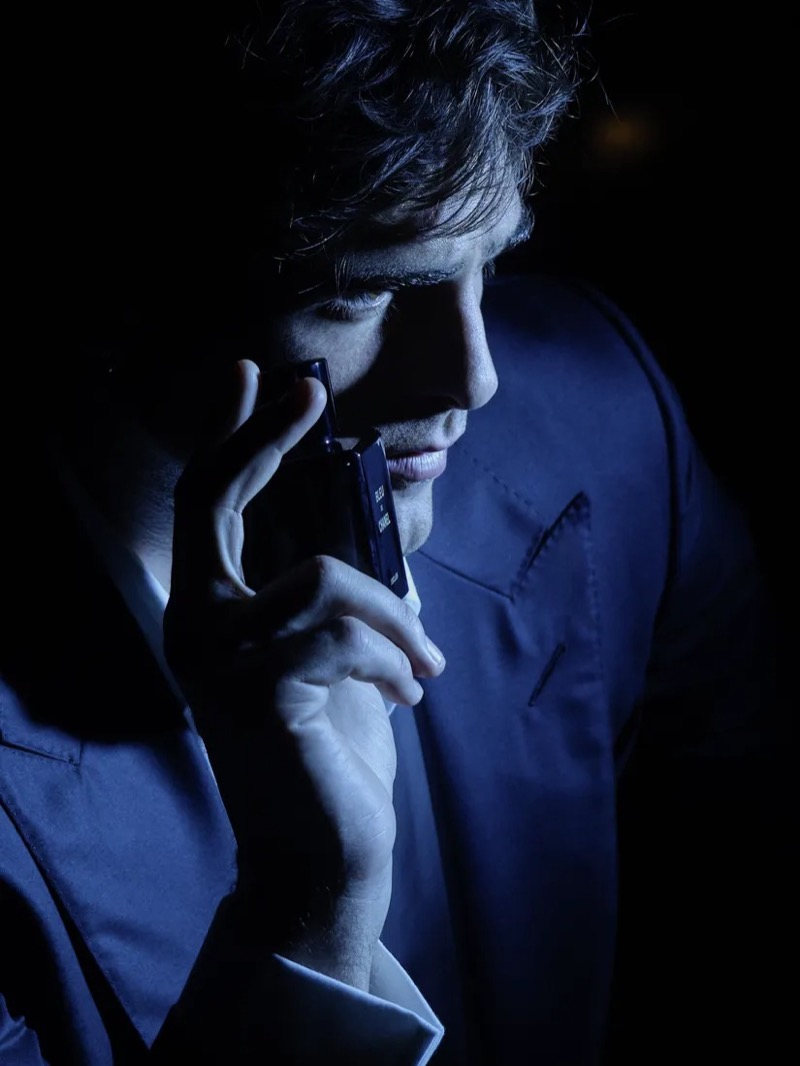 Elordi holds the Bleu de Chanel bottle to his cheek in a dark navy shirt and jacket under low blue light for the fragrance campaign