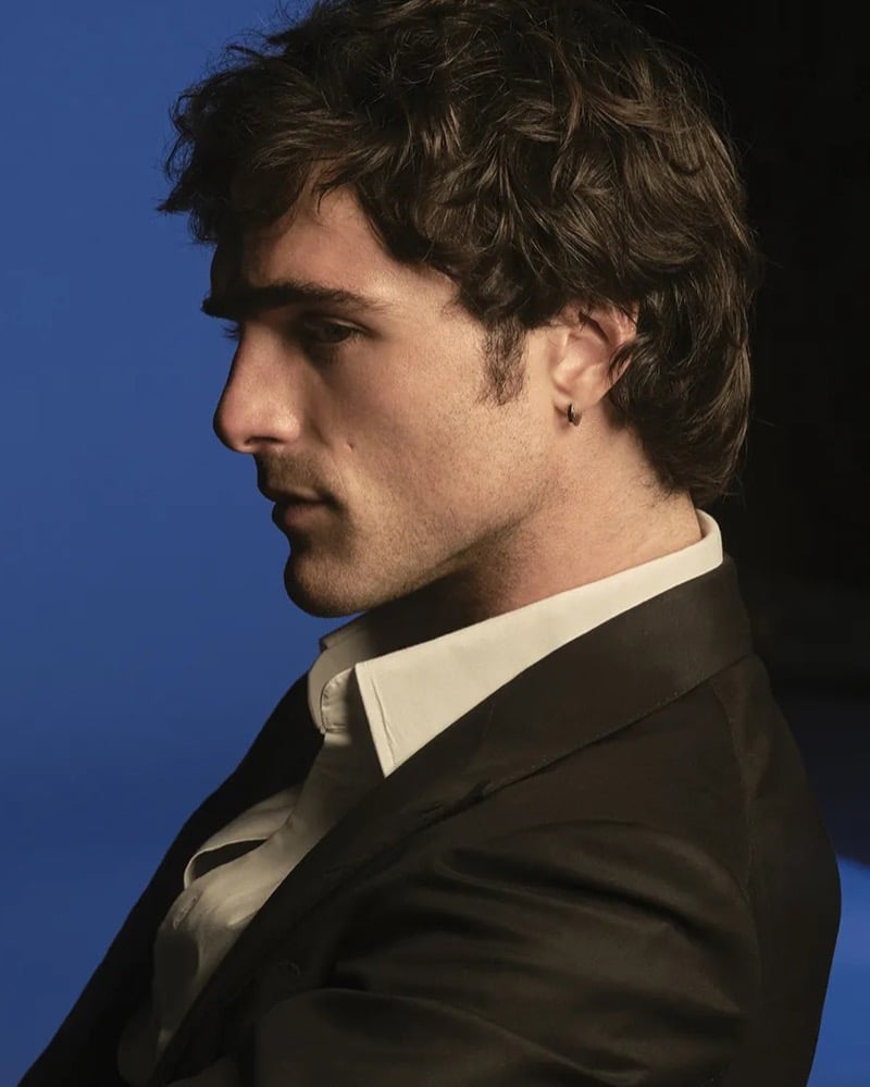 Elordi poses in profile against a deep blue backdrop in a dark suit jacket and open white shirt with a small hoop earring for Bleu de Chanel