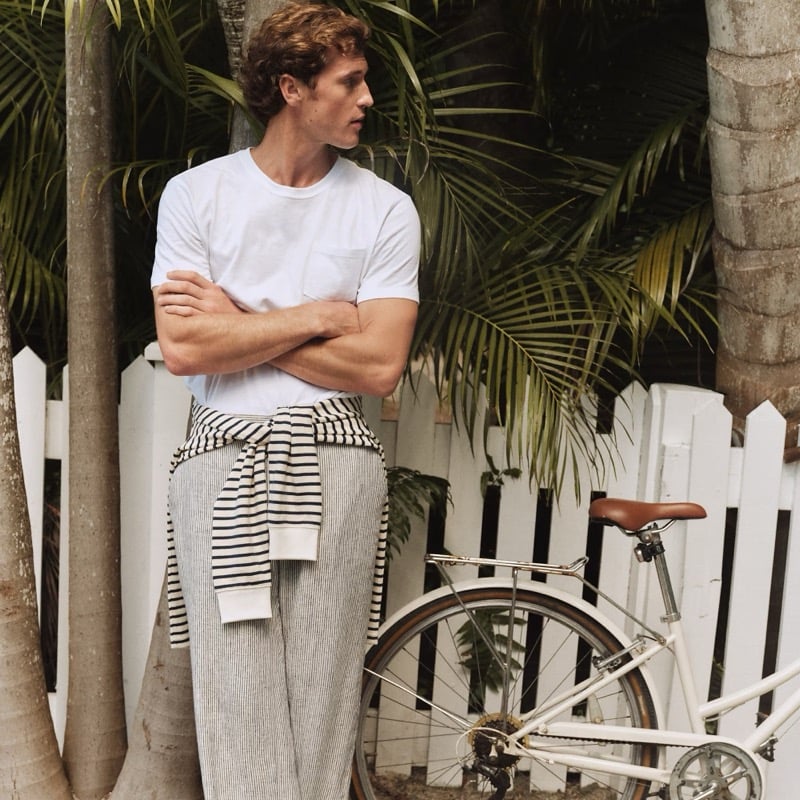 Jacques stands against a white picket fence in a white pocket tee, striped sweater tied at the waist, and gray linen trousers