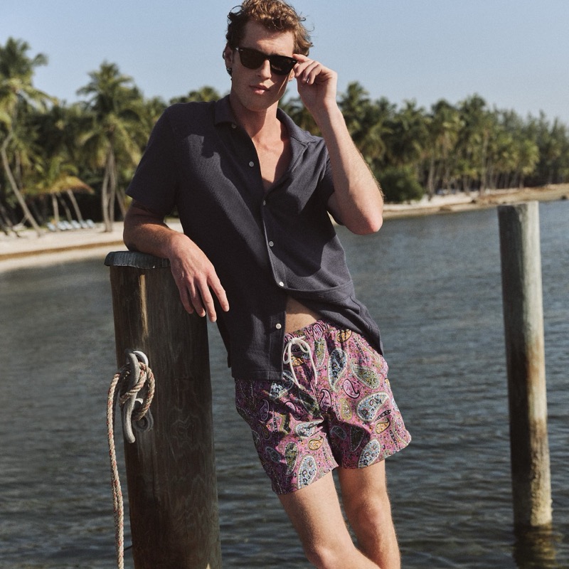 Jacques stands on a dock in sunglasses, a navy textured camp collar shirt, and paisley-print swim trunks