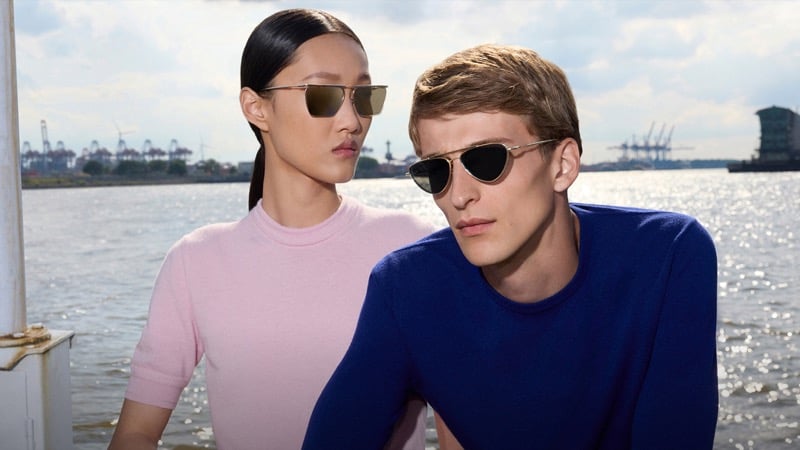 Feng and Vasko wear Jil Sander x Oliver Peoples titanium sunglasses in Hamburg harbor — she in slim rectangular frames, he in slim pilot frames