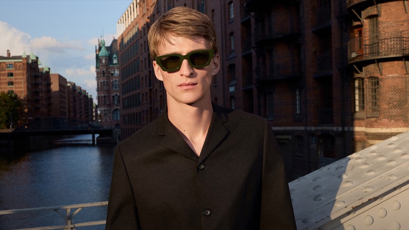 Vasko wears Jil Sander x Oliver Peoples Edition 6 sunglasses in dark green acetate against the brick warehouses of Hamburg's Speicherstadt