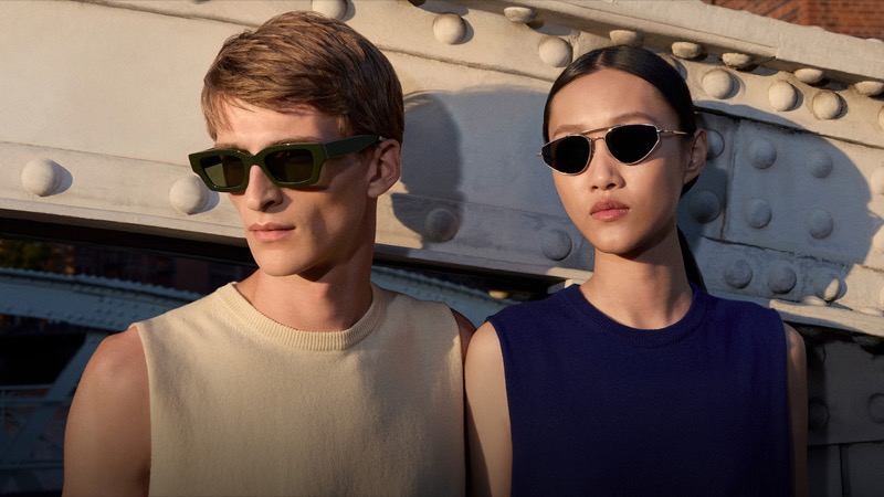 Vasko wears Jil Sander x Oliver Peoples Edition 6 sunglasses in dark green acetate alongside Feng in round titanium frames against a Hamburg bridge