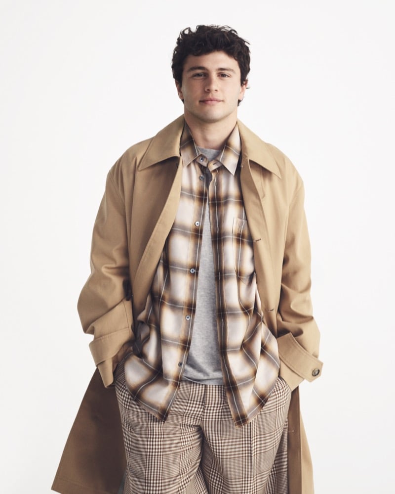 João Neves layers a camel Ami Paris trench coat over a brown plaid flannel and gray tee with brown houndstooth trousers