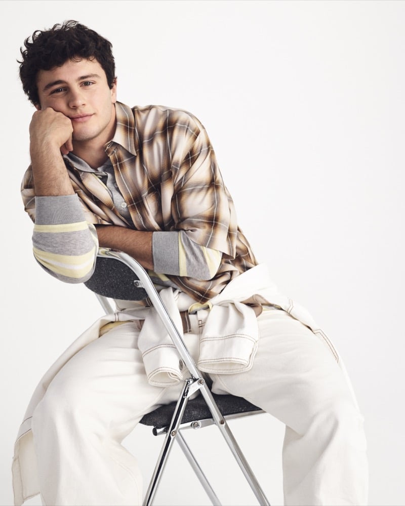 João Neves seated on a metal folding chair in a tan plaid flannel over a gray and yellow striped long-sleeve with white denim trousers