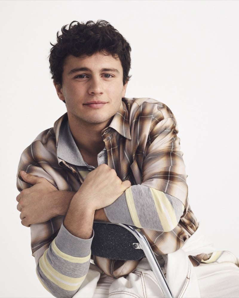 João Neves crosses his arms over the back of a folding chair in a tan plaid Ami Paris flannel with gray and yellow striped jersey sleeves visible underneath