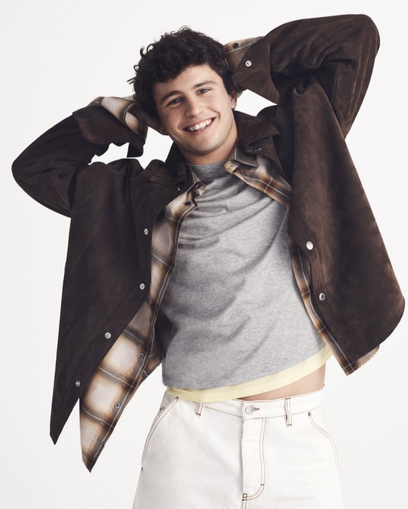 João Neves smiles with both hands behind his head in a brown suede overshirt over a plaid flannel, gray tee, yellow undershirt, and cream jeans
