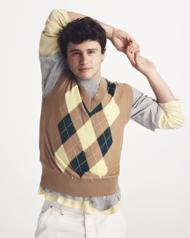 João Neves wears a camel, yellow, and fir green argyle knit vest layered over a gray tee and pale yellow long-sleeve with cream trousers